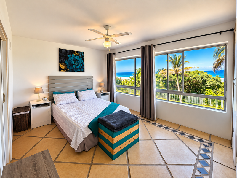Spacious bedroom featuring tiled flooring, a ceiling fan, and large sliding windows offering scenic ocean and garden views.