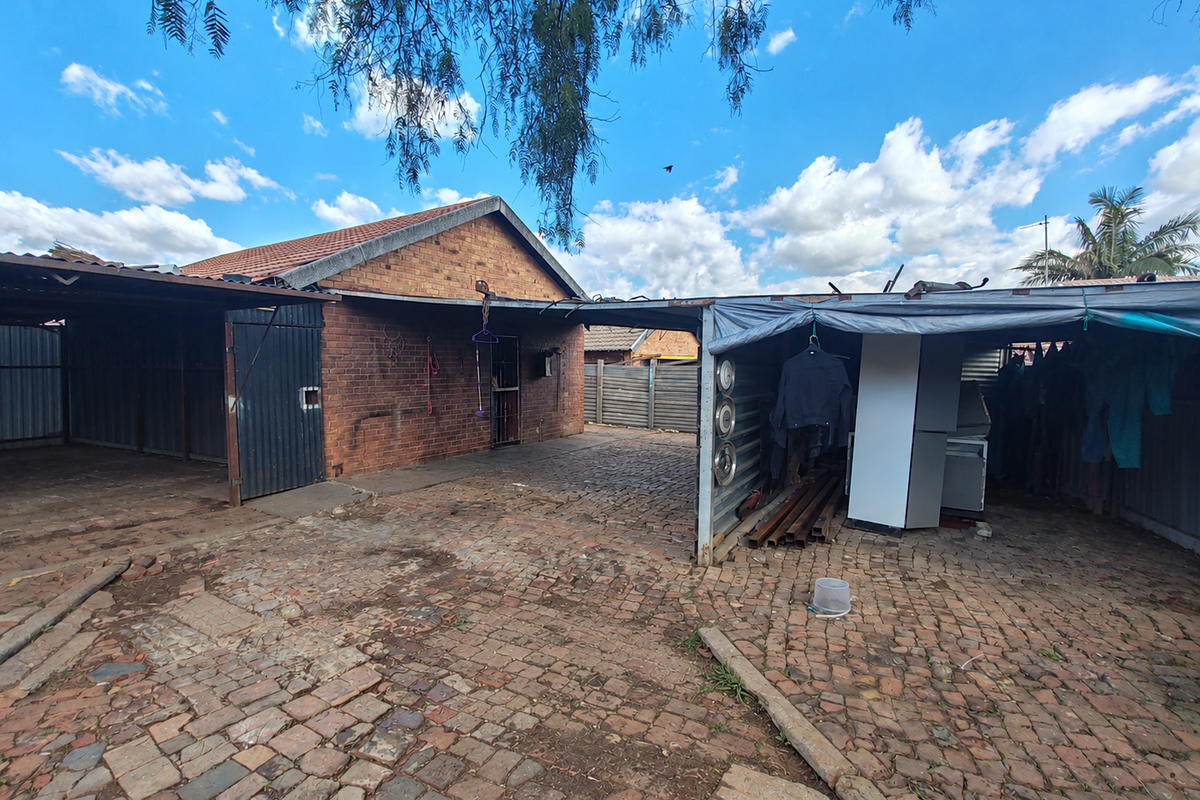 Rear exterior area featuring a brick house facade, paved driveway, and metal carport structures in need of maintenance.