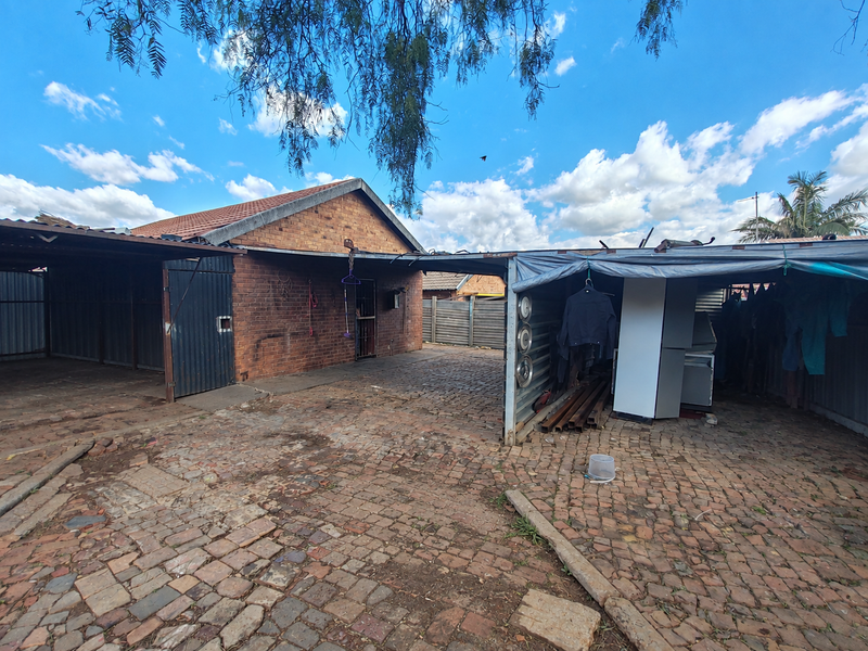Rear exterior area featuring a brick house facade, paved driveway, and metal carport structures in need of maintenance.