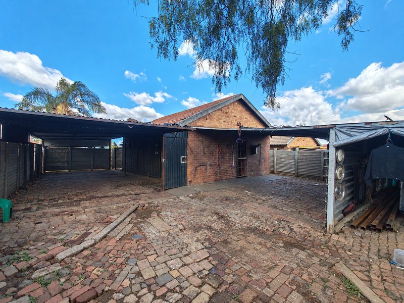 Rear exterior view of the property featuring a brick facade, tiled roof, and extensive paved driveway area with integrated carport structures.