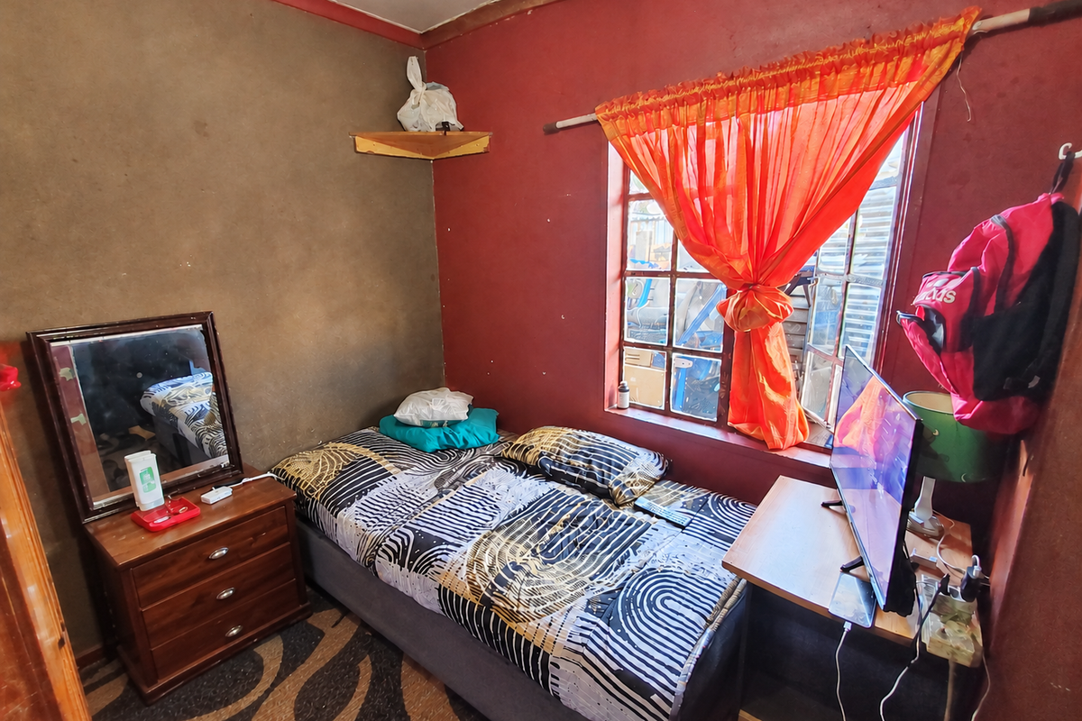 A compact bedroom featuring a window with a curtain rod and painted walls showing signs of wear and surface deterioration.