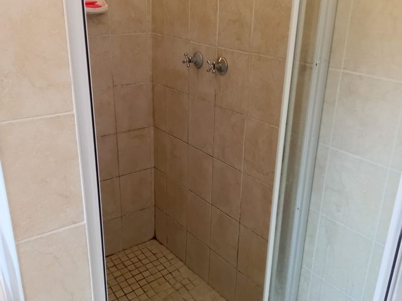 A compact shower stall featuring beige wall tiles, a tiled floor, and a standard shower mixer, currently showing signs of wear and grout discoloration.