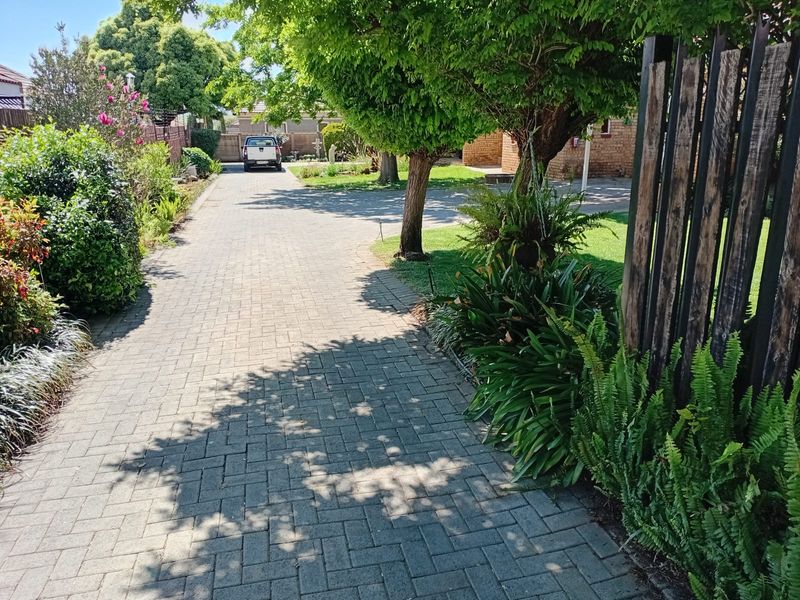 A well-maintained paved driveway leading to the property entrance, flanked by established trees, lush garden beds, and a timber boundary fence.