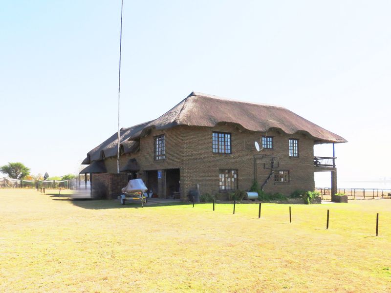 A rear view of a two-story brick house featuring a traditional thatch roof, grid windows, and an attached balcony overlooking the surrounding grounds.