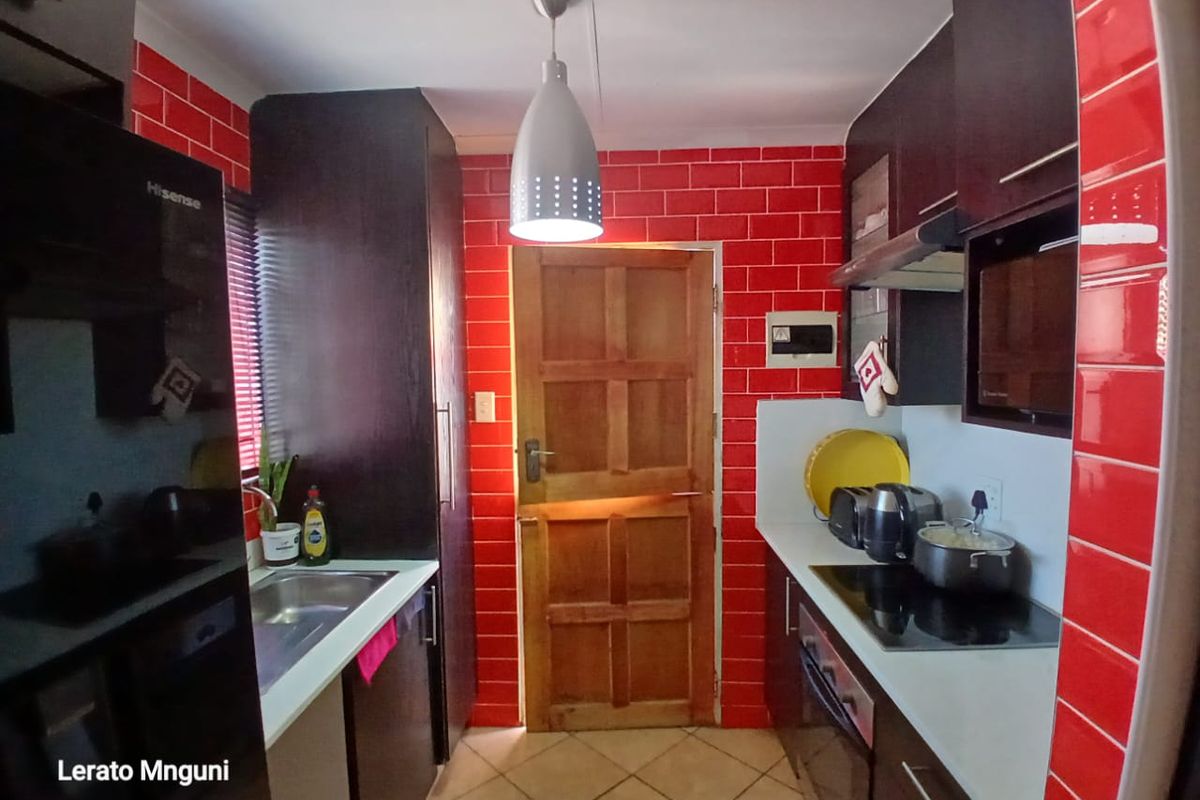 A compact kitchen featuring dark cabinetry, a red tiled splashback, electric hob, built-in oven, and tiled flooring with direct access to an exterior door.