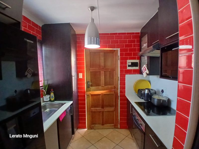 A compact kitchen featuring dark cabinetry, a red tiled splashback, electric hob, built-in oven, and tiled flooring with direct access to an exterior door.