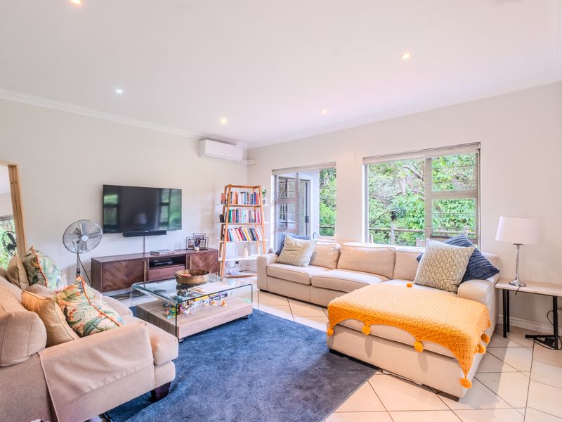A bright and spacious living room featuring tiled flooring, recessed downlighting, split-system air conditioning, and large windows providing natural light and garden views.