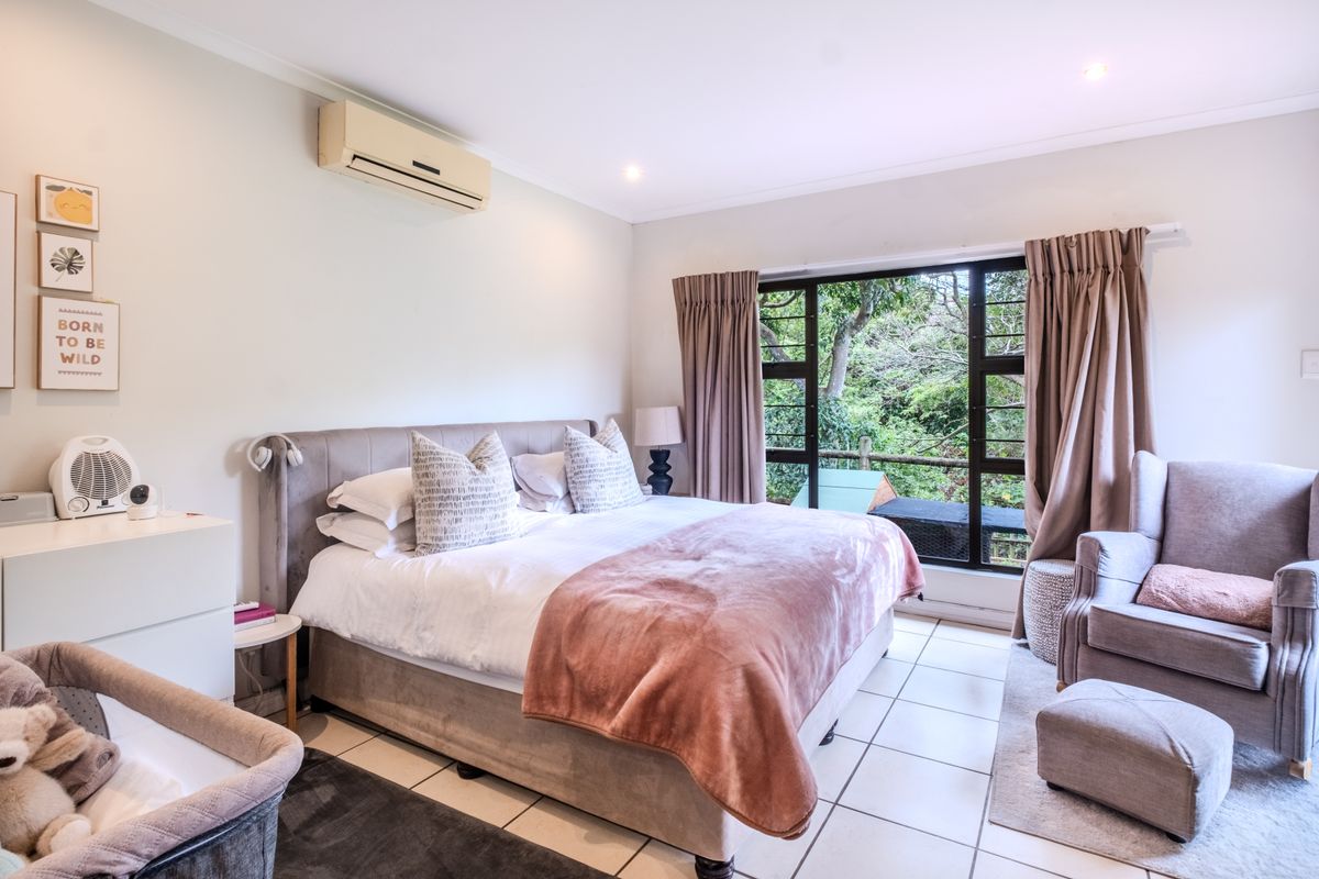 A well-lit bedroom featuring tiled flooring, a wall-mounted split-system air conditioner, and a large window offering a view of the surrounding greenery.