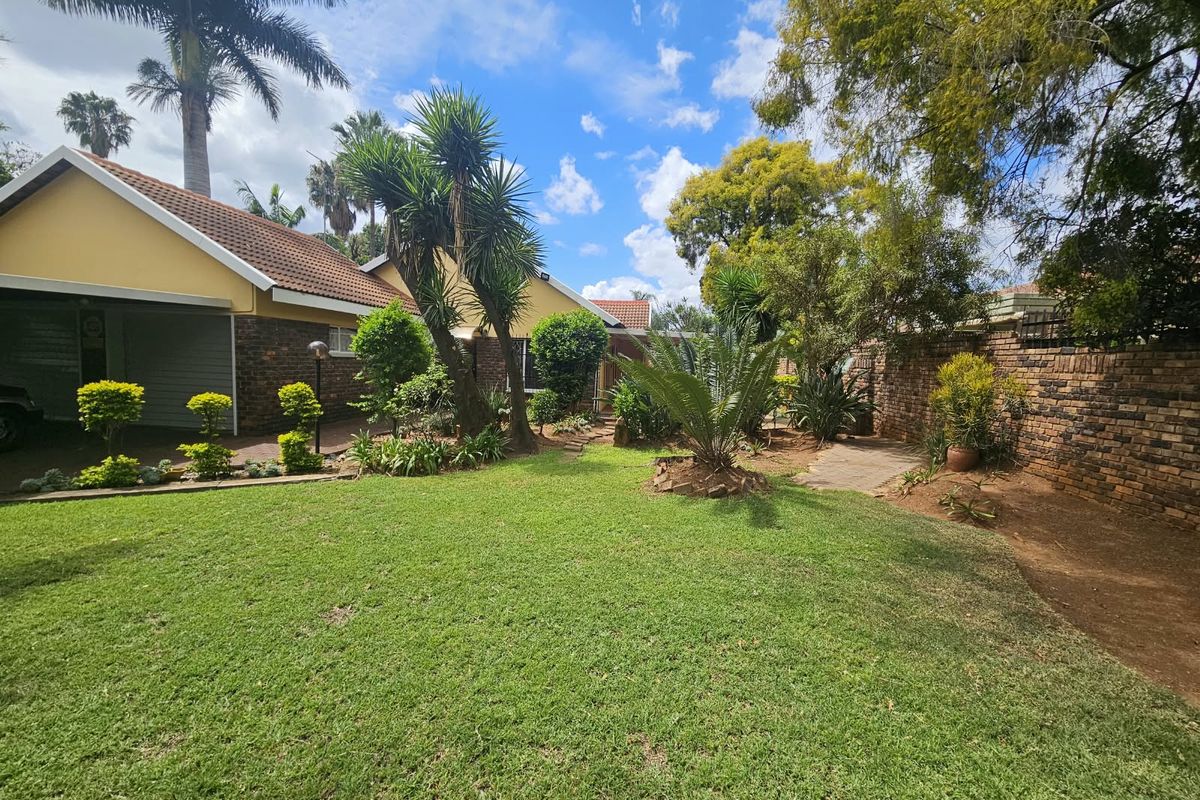 A well-maintained rear garden featuring a lush lawn, mature trees, and a brick boundary wall, adjacent to the property's garage and paved driveway.