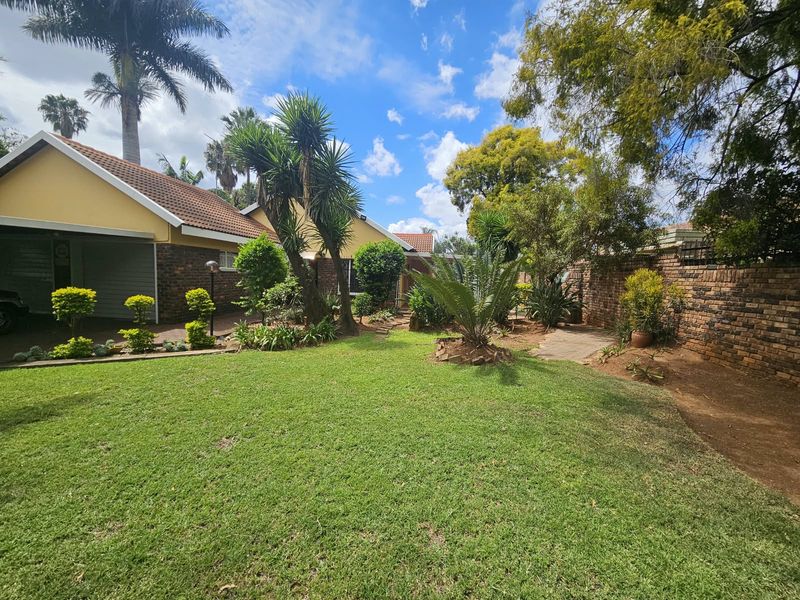 A well-maintained rear garden featuring a lush lawn, mature trees, and a brick boundary wall, adjacent to the property's garage and paved driveway.