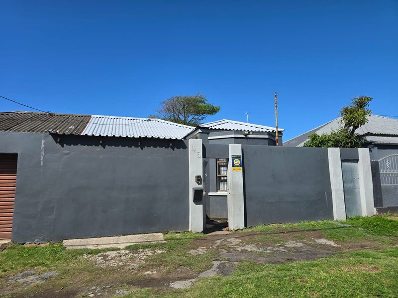 Front exterior view of the property featuring a grey boundary wall, pedestrian entrance, and corrugated metal roofing under a clear blue sky.