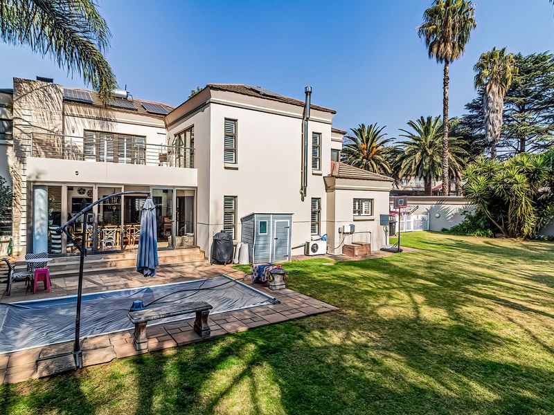 Spacious rear garden featuring a swimming pool with cover, paved patio area, and a two-story residence with balcony and solar panels.