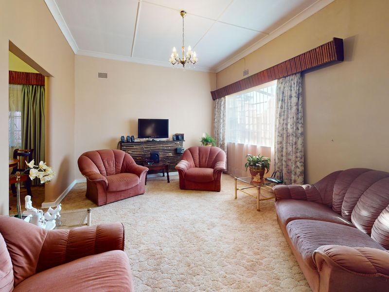 A spacious living room featuring neutral wall tones, a classic ceiling design, and large windows with security bars and decorative pelmets.