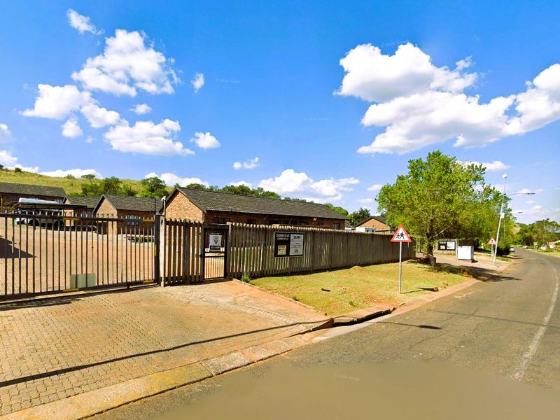 Street view of the townhouse complex entrance featuring a paved driveway, secure metal gate, and timber perimeter fencing.