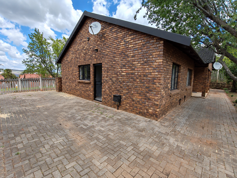 A well-maintained face brick townhouse exterior featuring a paved driveway, secure entrance, and tiled roof, set in a neat residential environment.