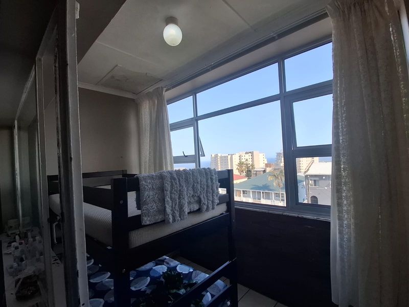 A bedroom featuring large aluminium-framed windows with urban views, tiled flooring, and a ceiling light fixture, requiring some cosmetic updates.
