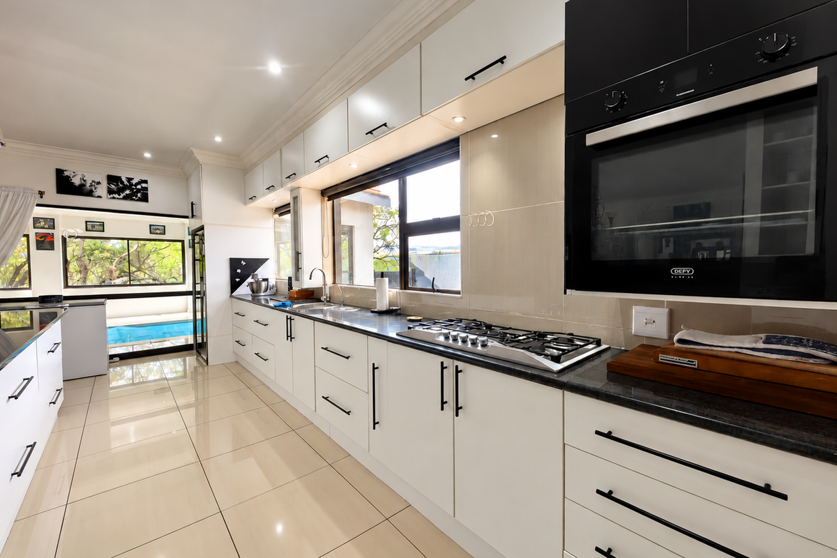 Modern kitchen featuring sleek white cabinetry, stone countertops, built-in oven, gas hob, and polished tiled flooring with a view towards the pool area.