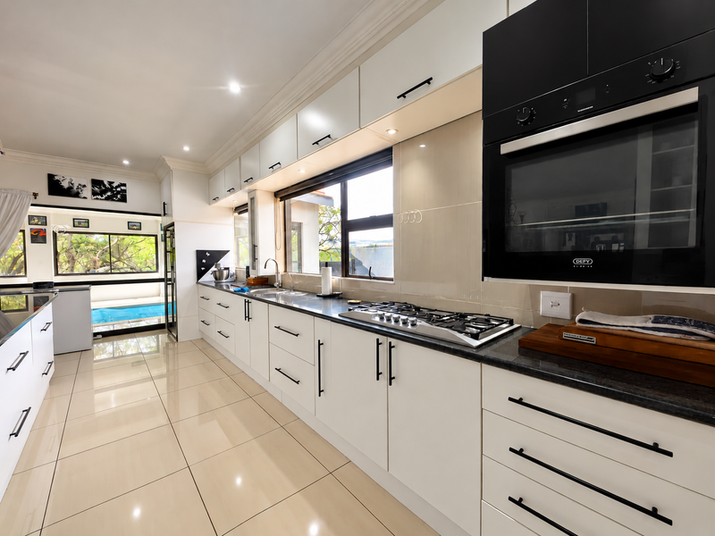 Modern kitchen featuring sleek white cabinetry, stone countertops, built-in oven, gas hob, and polished tiled flooring with a view towards the pool area.