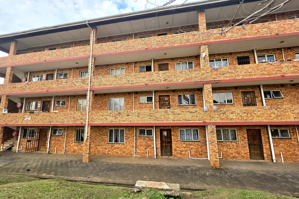 A multi-story apartment building exterior featuring face brick construction, paved ground level access, and security gates on individual unit entrances.