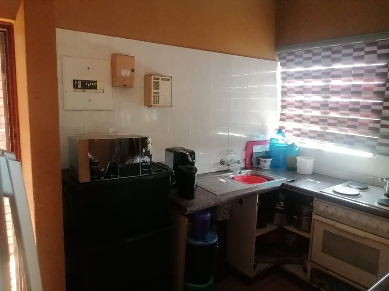 A compact kitchen area featuring a built-in oven, electric hob, stainless steel sink, and tiled splashback, with visible electrical distribution board components.