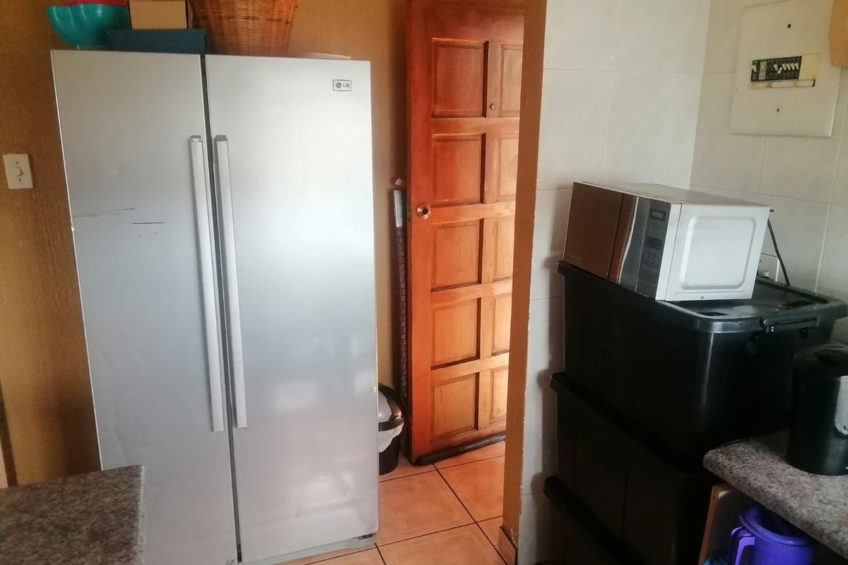 A kitchen area featuring tiled flooring, a timber door, and an exposed electrical distribution board, currently used for storage and appliance placement.