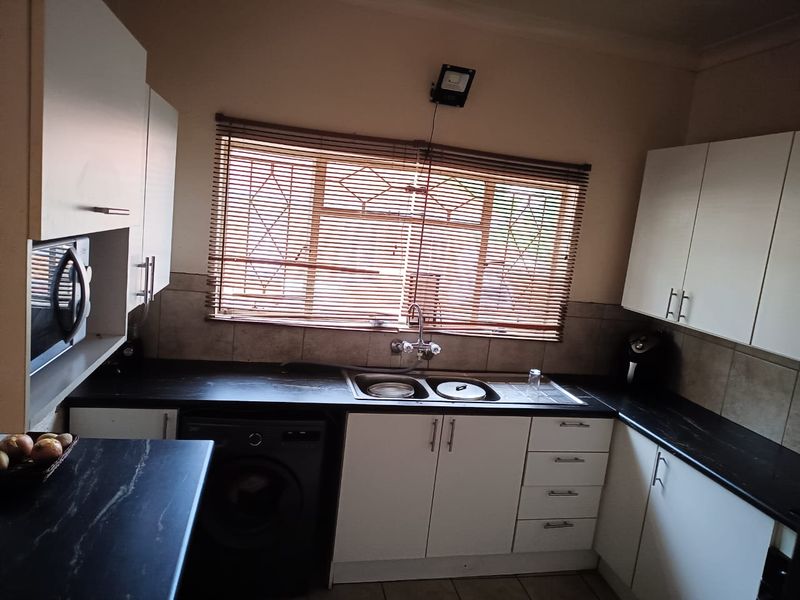 Functional kitchen featuring white cabinetry, dark laminate countertops, double sink, and integrated laundry appliance with tiled backsplash and window blinds.