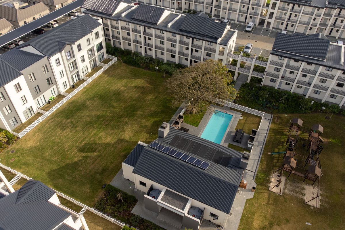 Aerial view of a modern apartment complex featuring a communal swimming pool, landscaped gardens, and residential buildings with solar-equipped metal roofs.