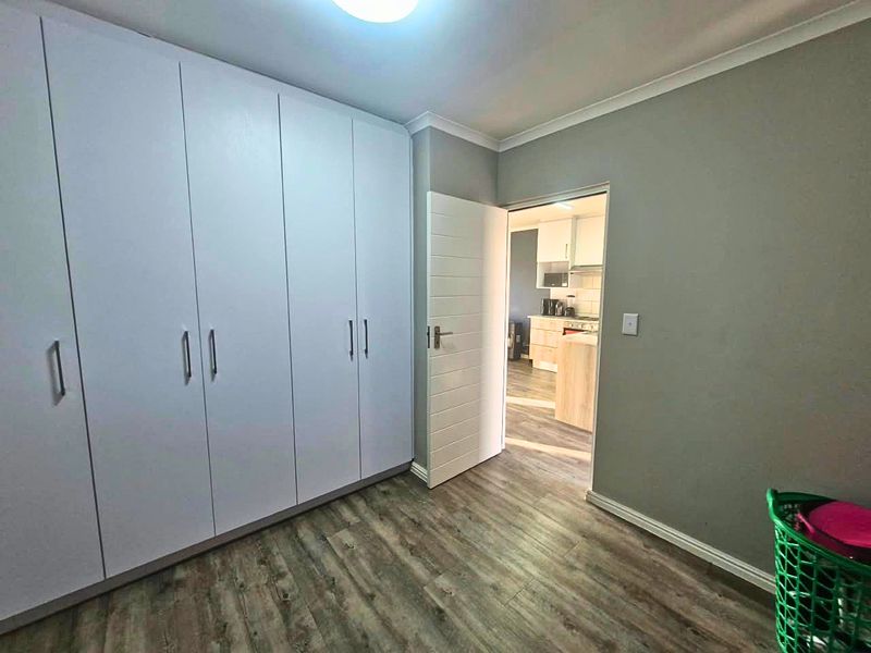 A clean, empty bedroom featuring modern white built-in cupboards, durable laminate flooring, and neutral grey walls, with a view into the adjacent kitchen.