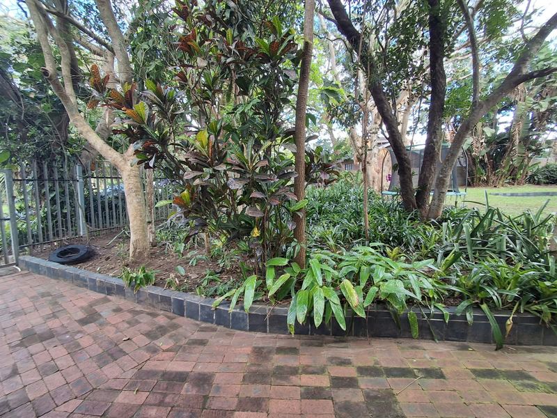 A lush garden area featuring mature trees, a raised garden bed, and a brick-paved patio area enclosed by metal perimeter fencing.