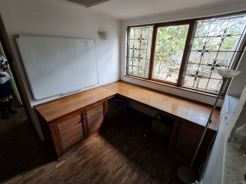 A functional home office featuring a large built-in wooden desk with integrated storage cabinetry, parquet flooring, and large windows with security bars.