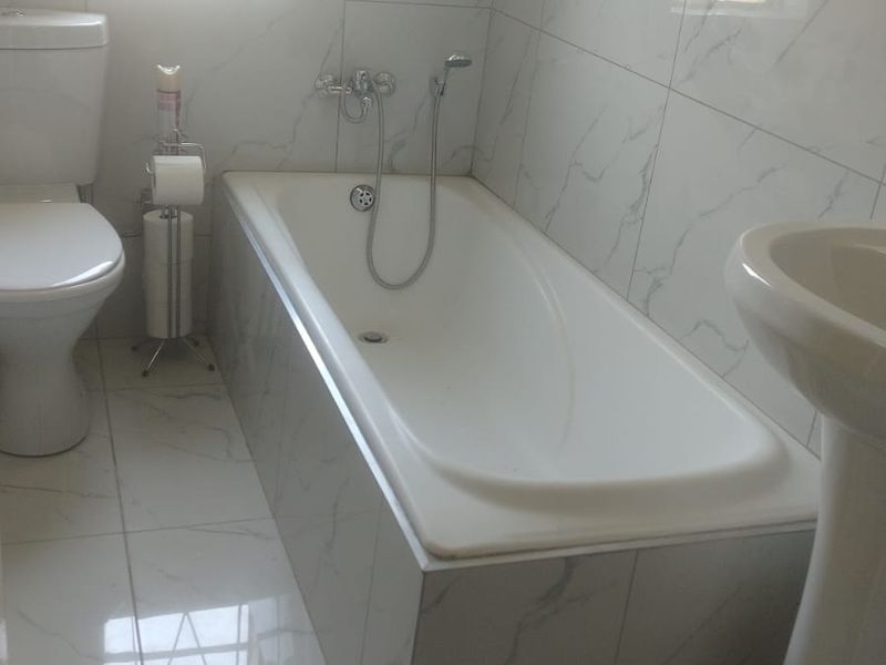 A clean, modern bathroom featuring white marble-look tiled walls and floors, a built-in bathtub, a pedestal basin, and a standard toilet.