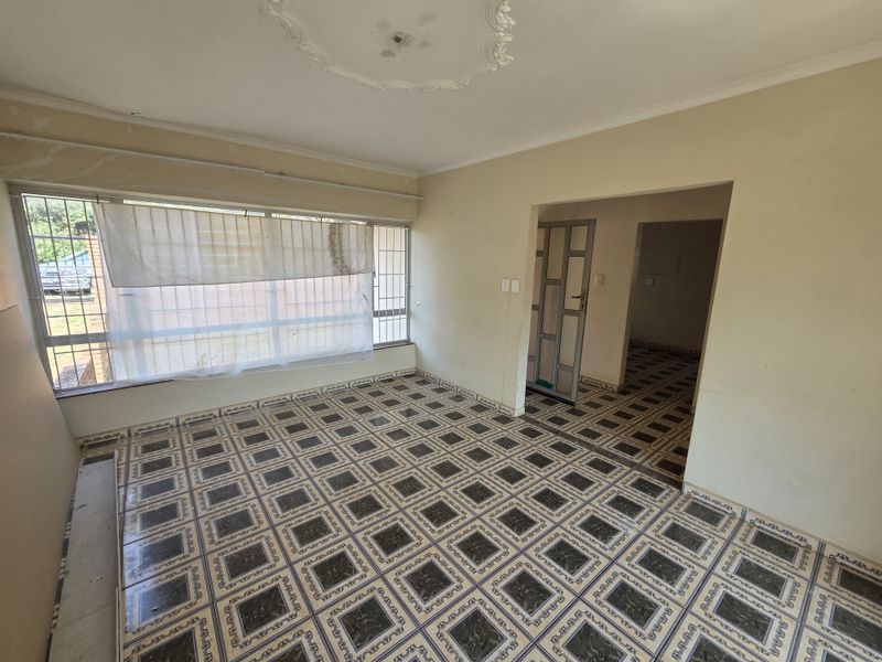 An empty living room featuring patterned tiled flooring, a decorative ceiling rose, and large windows, presenting a blank canvas for renovation.