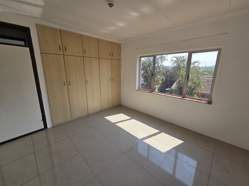 A bright, empty bedroom featuring neutral tiled flooring, ample built-in cupboard storage, and large windows providing natural light and a pleasant outlook.