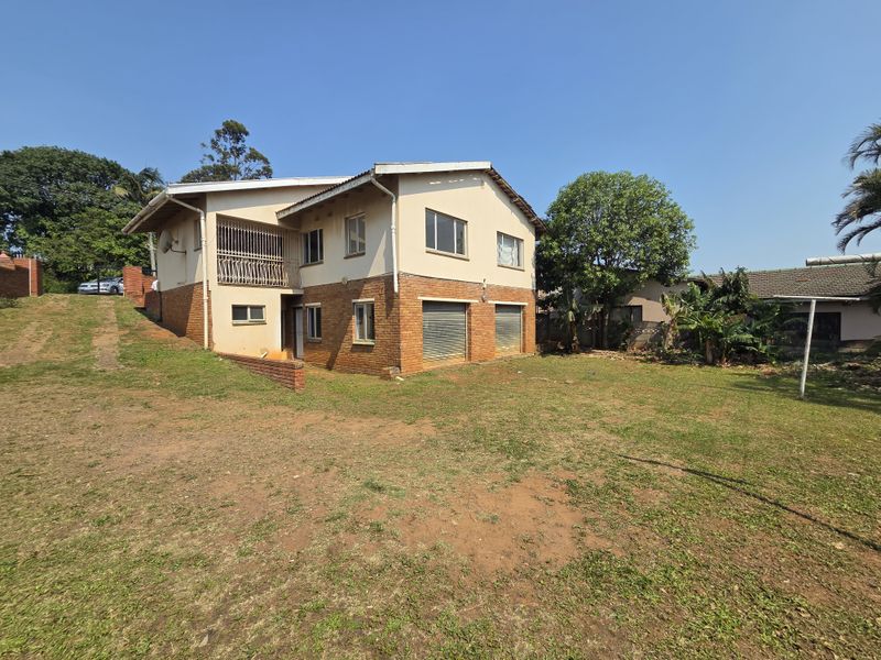 Rear view of a two-story house featuring a combination of brick and plastered walls, security shutters, and an elevated balcony overlooking a large garden.