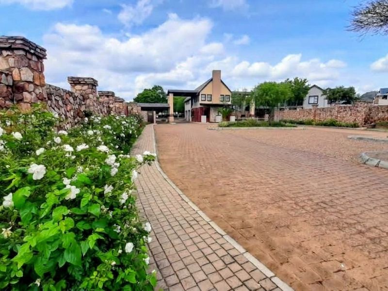 A well-maintained residential estate entrance featuring a stone boundary wall, paved driveway, and manicured garden landscaping leading to the main gate.