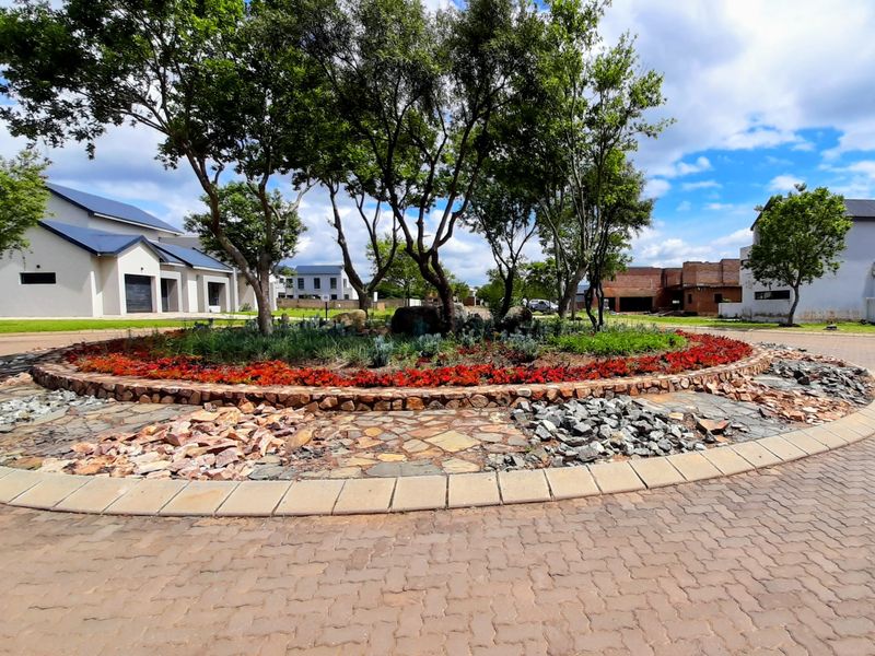 A well-maintained residential estate entrance featuring a landscaped roundabout with mature trees, stone detailing, and paved driveway access.