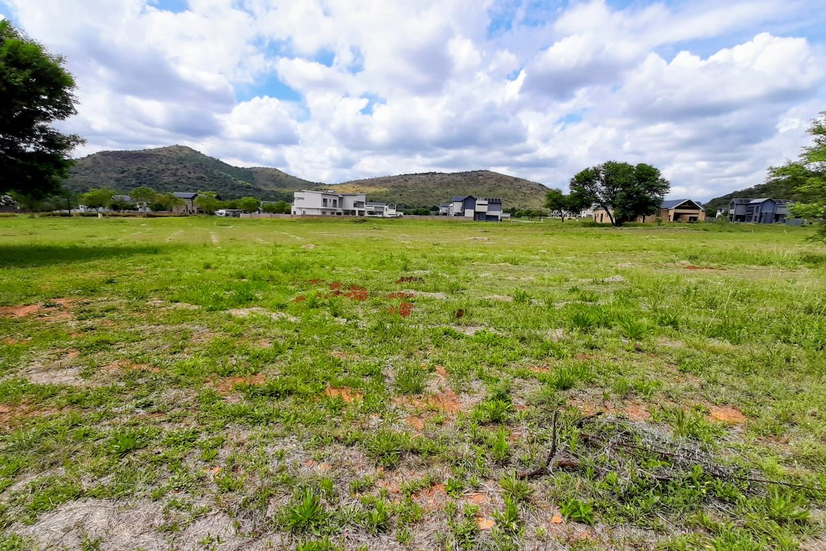 A scenic view of a vacant residential plot featuring natural grassland, mature trees, and a picturesque mountain backdrop in a developed estate.