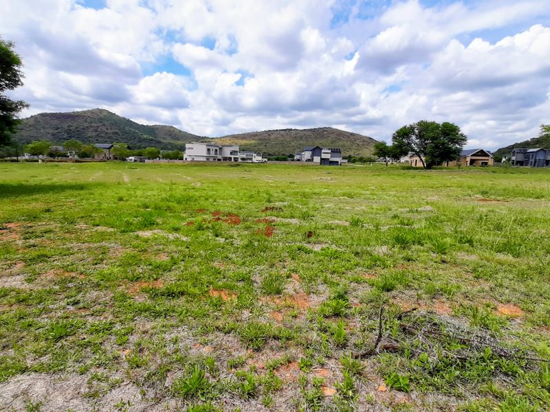 A scenic view of a vacant residential plot featuring natural grassland, mature trees, and a picturesque mountain backdrop in a developed estate.
