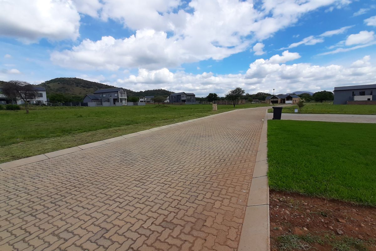 A view of a paved residential street within a developed estate, featuring manicured verges and surrounding vacant residential plots.