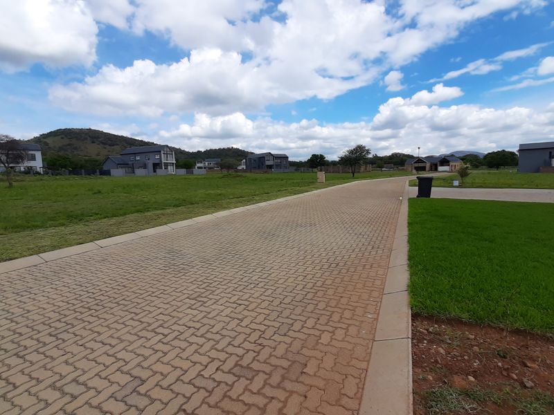 A view of a paved residential street within a developed estate, featuring manicured verges and surrounding vacant residential plots.