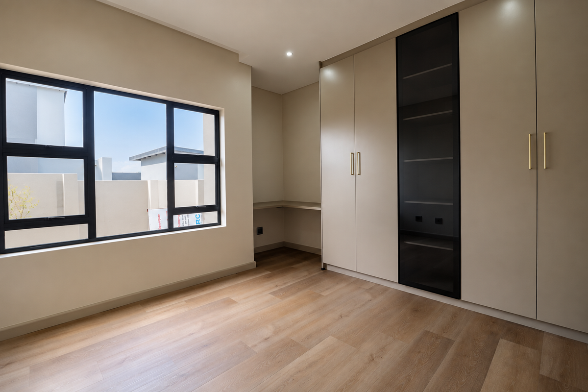 A modern, empty bedroom featuring light laminate flooring, built-in cupboards with a glass display section, and a built-in desk area under a large window.