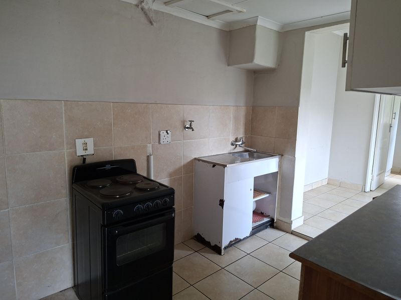 A functional kitchen area featuring a freestanding stove, stainless steel sink, tiled splashback, and ceramic floor tiles, currently in need of cosmetic updates.