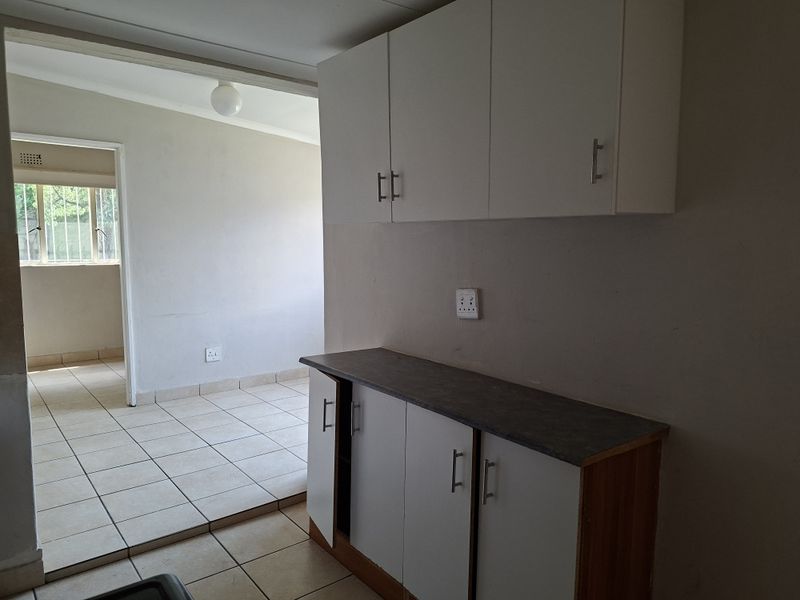 A functional kitchen area featuring built-in white cabinetry, laminate countertops, and ceramic floor tiles, leading into an adjacent living space.