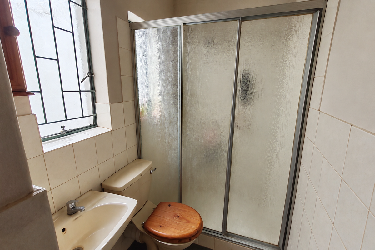 A compact bathroom featuring a shower enclosure, pedestal basin, and toilet, finished with neutral wall tiles and security bars on the window.