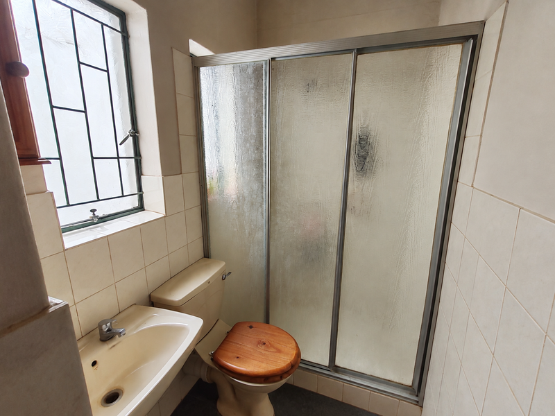 A compact bathroom featuring a shower enclosure, pedestal basin, and toilet, finished with neutral wall tiles and security bars on the window.