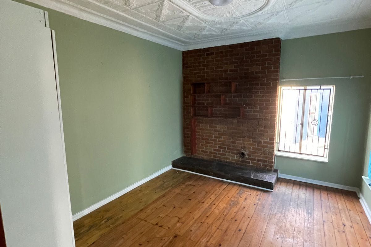 An empty living room featuring original pressed metal ceilings, a brick fireplace, and timber flooring, offering a character-filled space ready for renovation.