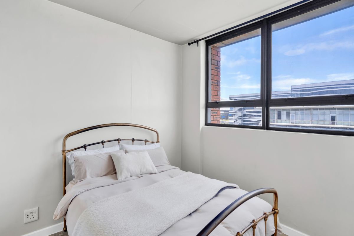 A bright bedroom featuring a large window with urban views and exposed brick detailing, offering a clean and neutral living space.
