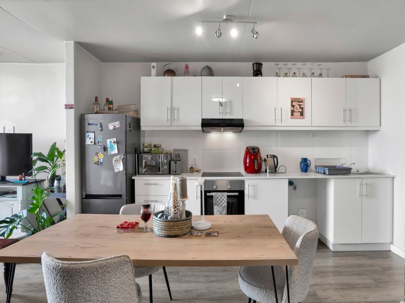 An open-plan kitchen and dining area featuring white cabinetry, integrated appliances, laminate flooring, and a distinctive exposed brick feature wall.