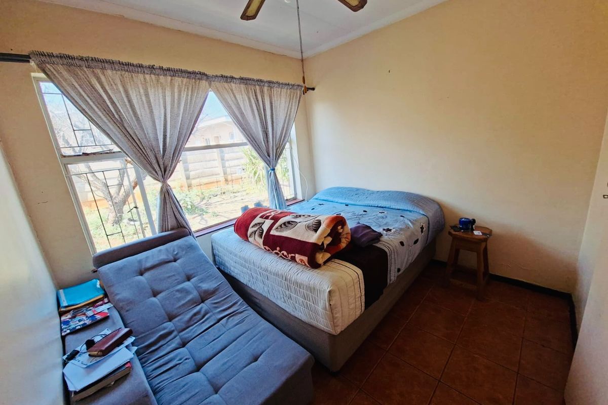 A bedroom featuring tiled flooring, a ceiling fan, and a large window with security bars, offering a functional space in need of cosmetic updates.