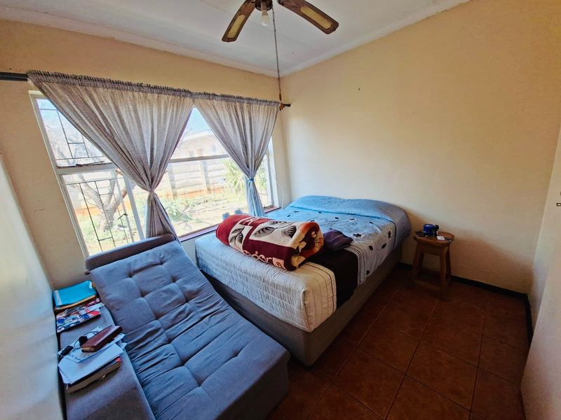 A bedroom featuring tiled flooring, a ceiling fan, and a large window with security bars, offering a functional space in need of cosmetic updates.
